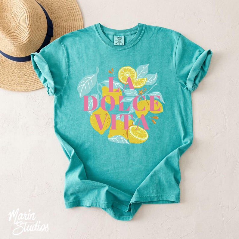 May include: A turquoise t-shirt featuring a lemon-themed graphic with the text "LA DOLCE VITA" in pink. The design includes illustrations of lemons, leaves, and floral accents. A straw hat is partially visible in the background.