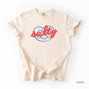 A flat lay of an Ivory Comfort Colors shirt with a design featuring a pretzel and the phrase Salty over the top of it.