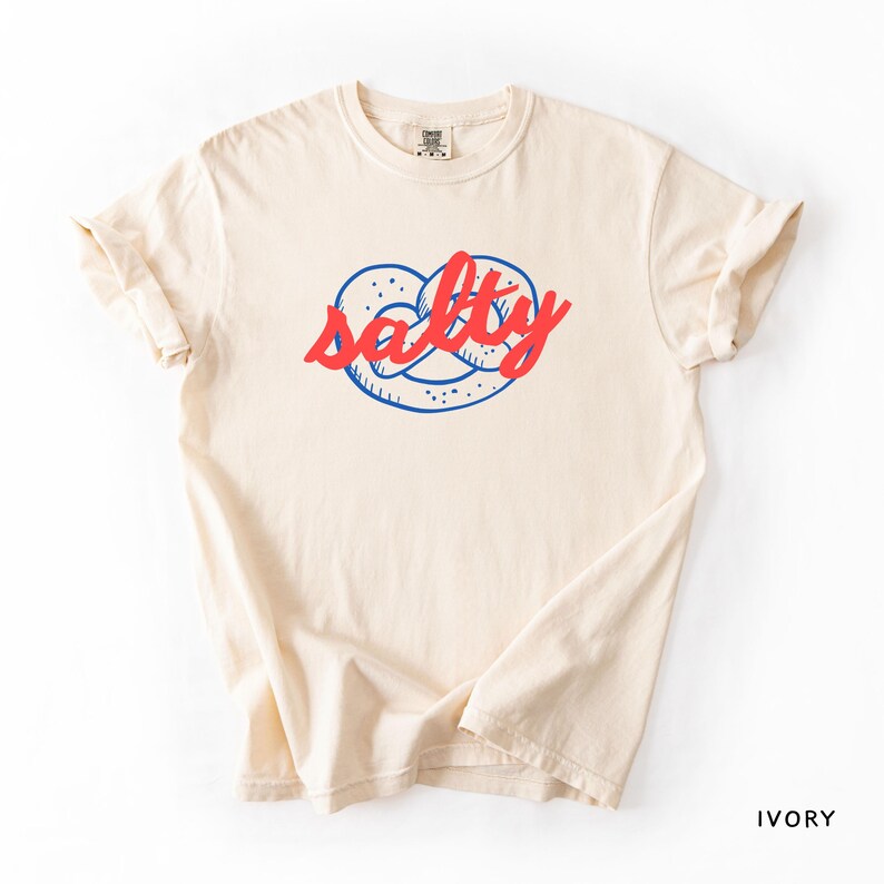 A flat lay of an Ivory Comfort Colors shirt with a design featuring a pretzel and the phrase Salty over the top of it.