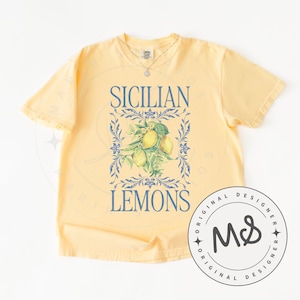 May include: Yellow t-shirt with the words "SICILIAN LEMONS" in blue, framed by a blue floral design. The graphic includes a cluster of lemons. A circular logo with "ORIGINAL DESIGNER" and "MS" is on the lower right.
