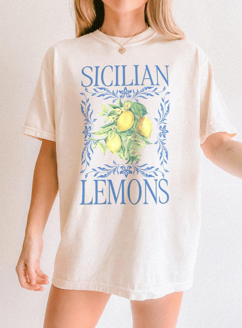 A woman is wearing an Ivory Comfort Colors shirt with a design featuring a branch of lemons and the phrase Sicilain Lemons.