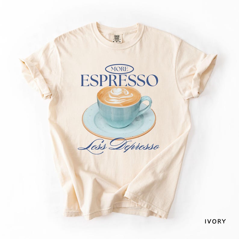 A flat lay of an Ivory Comfort Colors shirt with a design featuring an espresso and the phrase More Espresso Less Depresso.