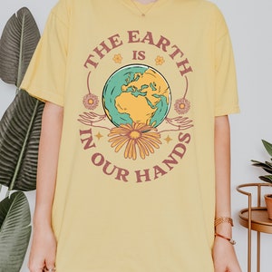 A woman is wearing a butter yellow Comfort Colors shirt with an Earth Day design that says The Earth is in Your Hands.