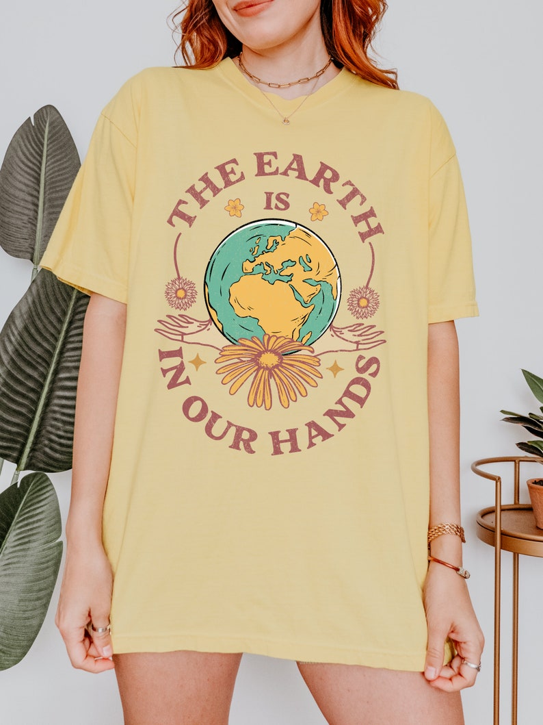 A woman is wearing a butter yellow Comfort Colors shirt with an Earth Day design that says The Earth is in Your Hands.