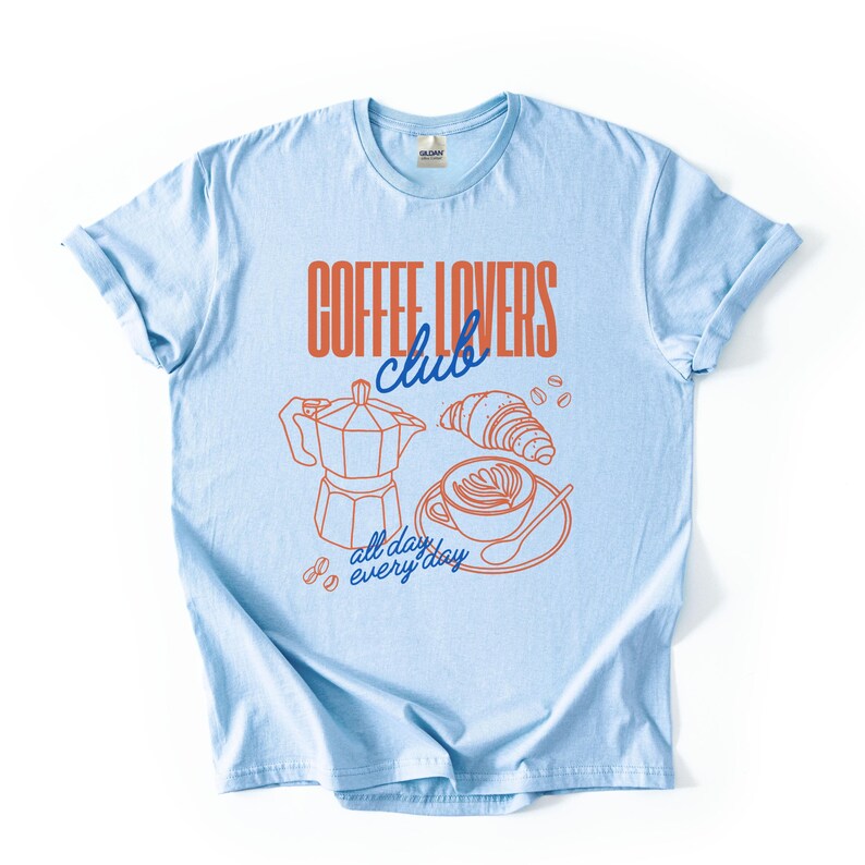 A  flat lay of a Light Blue Gildan 2000 t-shirt with a design featuring a hand-drawn coffee and croissant design that reads Coffee Lover Club, All Day Every Day.