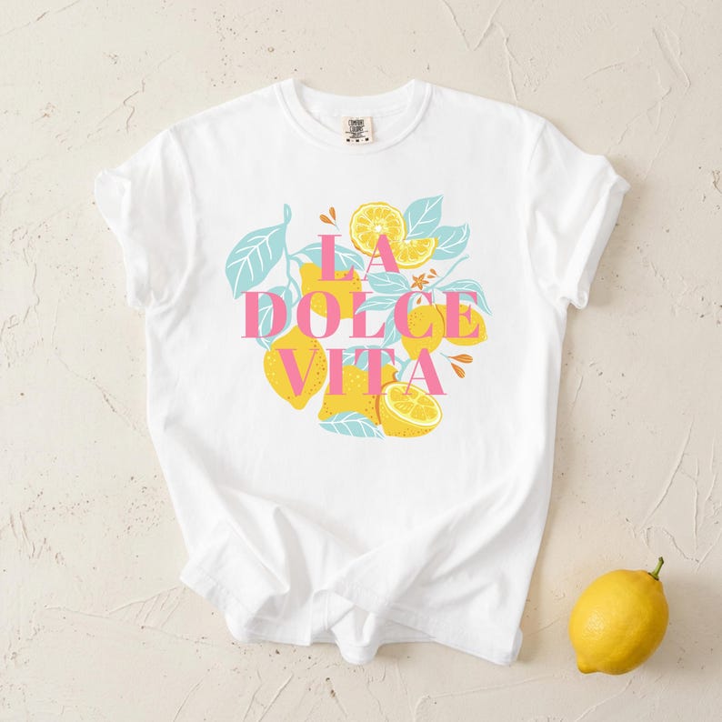 May include: White t-shirt with the text "LA DOLCE VITA" in pink, surrounded by yellow lemons and blue leaves. A single lemon rests on the surface. The shirt is a casual, comfortable style.