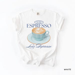 A flat lay of a white Comfort Colors shirt with a design featuring an espresso and the phrase More Espresso Less Depresso.