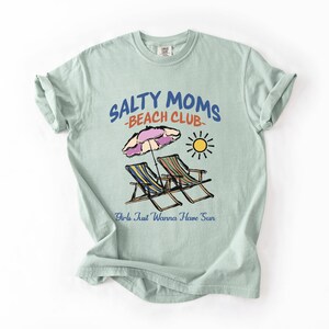 A Bay Comfort Colors shirt ona simple white background with a desgn  featuring a colorful hand-drawn scene of a striped beach umbrella and two chairs with the phrase Salty Moms Beach Club at the top and Girls Just Wanna Have Fun at the bottom of the design.