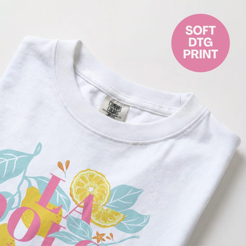 May include: White t-shirt with a lemon-themed graphic. The design includes pink text, yellow lemons, and blue-green leaves. The shirt has a crew neck and a tag that says "Comfort Colors". A pink circle with "SOFT DTG PRINT" is in the upper right.