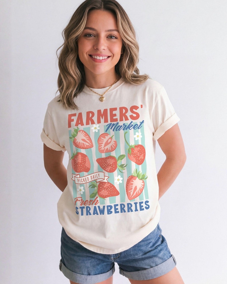 May include: Cream-colored t-shirt with a "FARMERS' MARKET" graphic featuring strawberries and the text "PICKED DAILY FRESH STRAWBERRIES". The design includes red strawberries, blue stripes, and white flowers. The shirt has rolled-up sleeves.