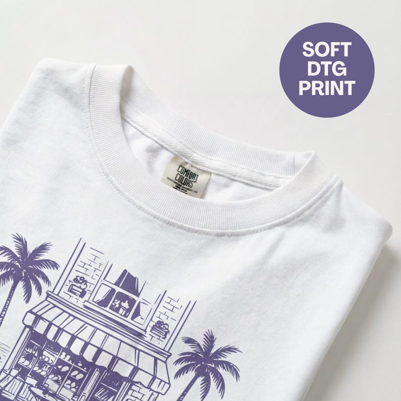 Peut inclure: T-shirt blanc avec une illustration violette d'une boutique et de palmiers. Le t-shirt a un col rond et une &eacute;tiquette "Comfort Colors". Un cercle violet avec "SOFT DTG PRINT" est en haut &agrave; droite.