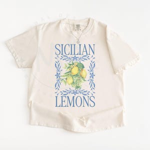 May include: Cream-colored t-shirt featuring the words "SICILIAN LEMONS" in blue, with a watercolor illustration of lemons and leaves. The design is framed by blue floral elements. A silver necklace is visible.
