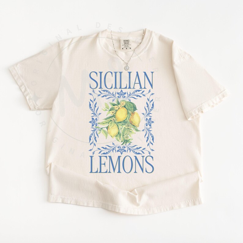 May include: Cream-colored t-shirt featuring the words "SICILIAN LEMONS" in blue, with a watercolor illustration of lemons and leaves. The design is framed by blue floral elements. A silver necklace is visible.