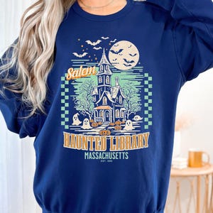 A woman is wearing an oversized Navy Comfort Colors sweatshirt with a design featuring a haunted library with ghosts and pumpkins and the phrase Salem Haunted Library, Massachusetts.