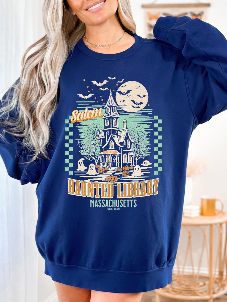 A woman is wearing an oversized Navy Comfort Colors sweatshirt with a design featuring a haunted library with ghosts and pumpkins and the phrase Salem Haunted Library, Massachusetts.