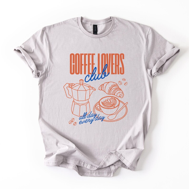 A  flat lay of an Ice Grey Gildan 2000 t-shirt with a design featuring a hand-drawn coffee and croissant design that reads Coffee Lover Club, All Day Every Day.