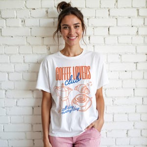 A woman is wearing a White 2000 t-shirt with a design featuring a hand-drawn coffee and croissant design that reads Coffee Lover Club, All Day Every Day.