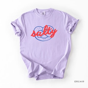 A flat lay of an Orchid Comfort Colors shirt with a design featuring a pretzel and the phrase Salty over the top of it.