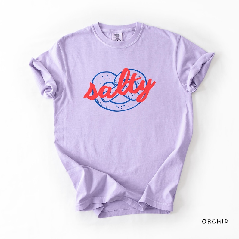 A flat lay of an Orchid Comfort Colors shirt with a design featuring a pretzel and the phrase Salty over the top of it.