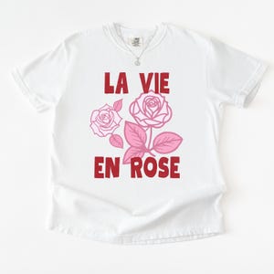 A flat lay of a white Comfort Colors t-shirt with bold red lettering that reads “La Vie En Rose.” The graphic features three outlined pink roses with stylized leaves, giving a vintage-inspired romantic look. She pairs the tee with pastel green gingham shorts for a soft, summery aesthetic.