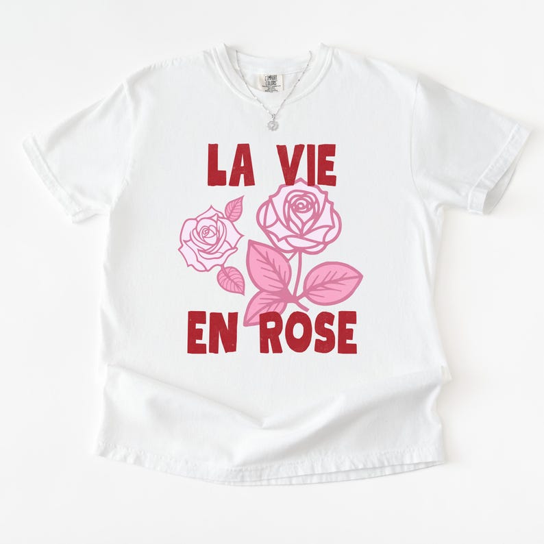 A flat lay of a white Comfort Colors t-shirt with bold red lettering that reads “La Vie En Rose.” The graphic features three outlined pink roses with stylized leaves, giving a vintage-inspired romantic look. She pairs the tee with pastel green gingham shorts for a soft, summery aesthetic.