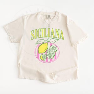 A flat lay of an Ivory Comfort Colors shirt with a design featuring a hand-drawn lemon and lemon slices on a plate, and the phrase Siciliana at the top in a serif font.