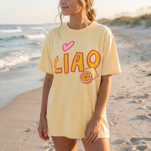 K&ouml;nnte beinhalten: Hellgelbes Kurzarm-T-Shirt mit dem Wort "CIAO" in orangefarbenen Buchstaben mit roter Umrandung, einem rosa Herz und einer Zitronenscheibe. Das Shirt wird am Strand getragen.