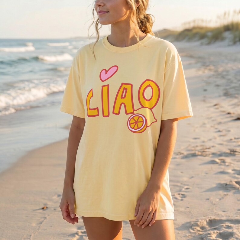 K&ouml;nnte beinhalten: Hellgelbes Kurzarm-T-Shirt mit dem Wort "CIAO" in orangefarbenen Buchstaben mit roter Umrandung, einem rosa Herz und einer Zitronenscheibe. Das Shirt wird am Strand getragen.