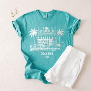 Peut inclure: Un t-shirt turquoise avec un dessin blanc d'un caf&eacute;, de palmiers et le texte "DOCKSIDE CAFE". Associ&eacute; &agrave; un short blanc et des boucles d'oreilles ancre dor&eacute;es, cet ensemble &eacute;voque une ambiance c&ocirc;ti&egrave;re d&eacute;contract&eacute;e.