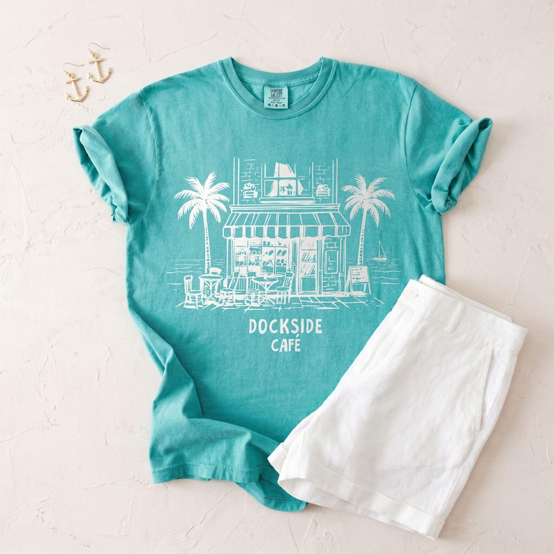 Peut inclure: Un t-shirt turquoise avec un dessin blanc d'un caf&eacute;, de palmiers et le texte "DOCKSIDE CAFE". Associ&eacute; &agrave; un short blanc et des boucles d'oreilles ancre dor&eacute;es, cet ensemble &eacute;voque une ambiance c&ocirc;ti&egrave;re d&eacute;contract&eacute;e.