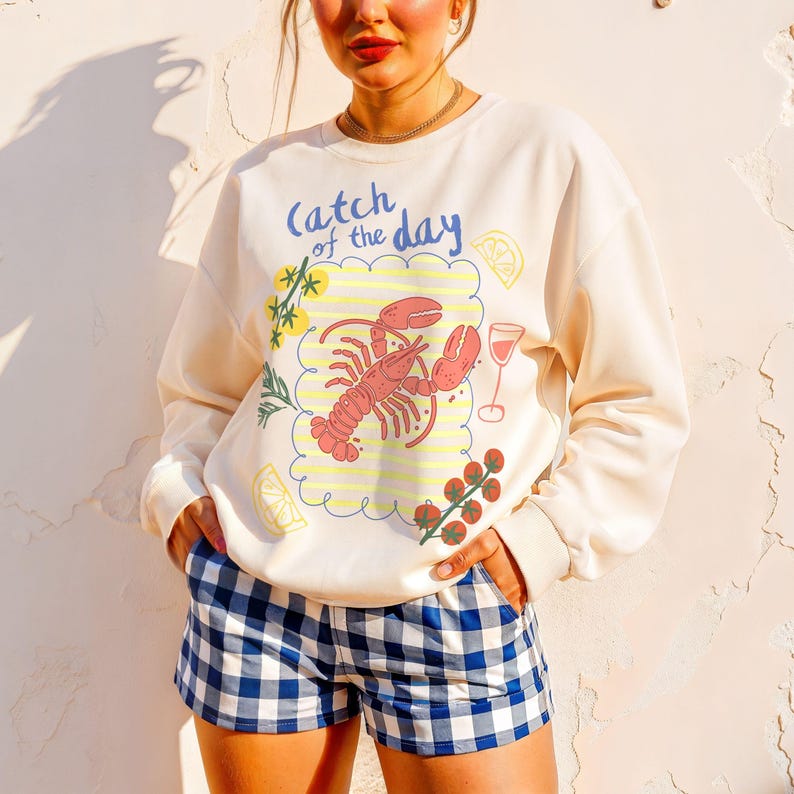 May include: Cream-colored sweatshirt with a lobster graphic and the text "Catch of the Day." The graphic includes a red lobster, a glass of wine, and lemon slices. The person is wearing blue and white checkered shorts.