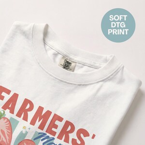 May include: White t-shirt with a crew neck featuring a graphic design. The design includes the words "FARMERS' Market" in red, along with illustrations of strawberries and other elements. The shirt also has a "SOFT DTG PRINT" label.