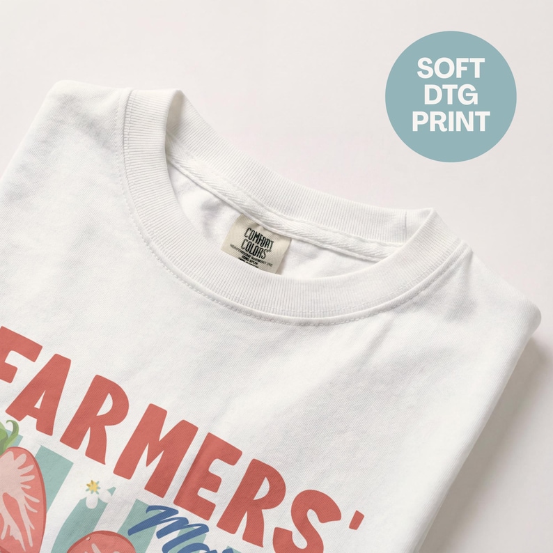 May include: White t-shirt with a crew neck featuring a graphic design. The design includes the words "FARMERS' Market" in red, along with illustrations of strawberries and other elements. The shirt also has a "SOFT DTG PRINT" label.