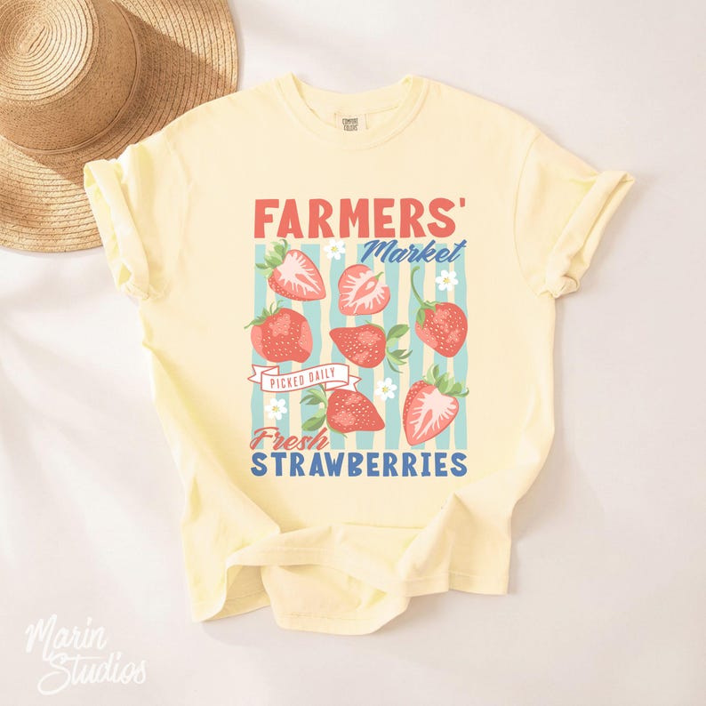 May include: A light yellow t-shirt with a graphic design featuring strawberries and the text "FARMERS' MARKET Fresh STRAWBERRIES". The design includes red strawberries, blue stripes, and white flowers.