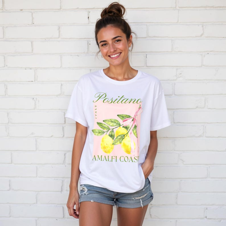A woman is wearing a white Gildan 2000 unisex T-shirt with a design featuring a branch of lemons with a long pink coquette bow tied around it and the phrase Positano, Amalfi Coast, Italy.