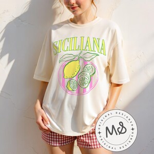A woman is wearing an ivory Comfort Colors shirt with a design featuring a hand-drawn lemon and lemon slices on a plate, and the phrase Siciliana at the top in a serif font.