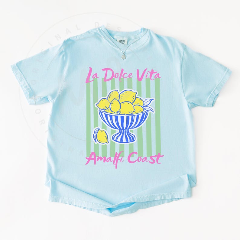 A flat lay of a Chambray Comfort Colors shirt with a. design featuring a bow of lemons against a striped background and the phrase La Dolce Vita, Amalfi Coast in a playful script font.