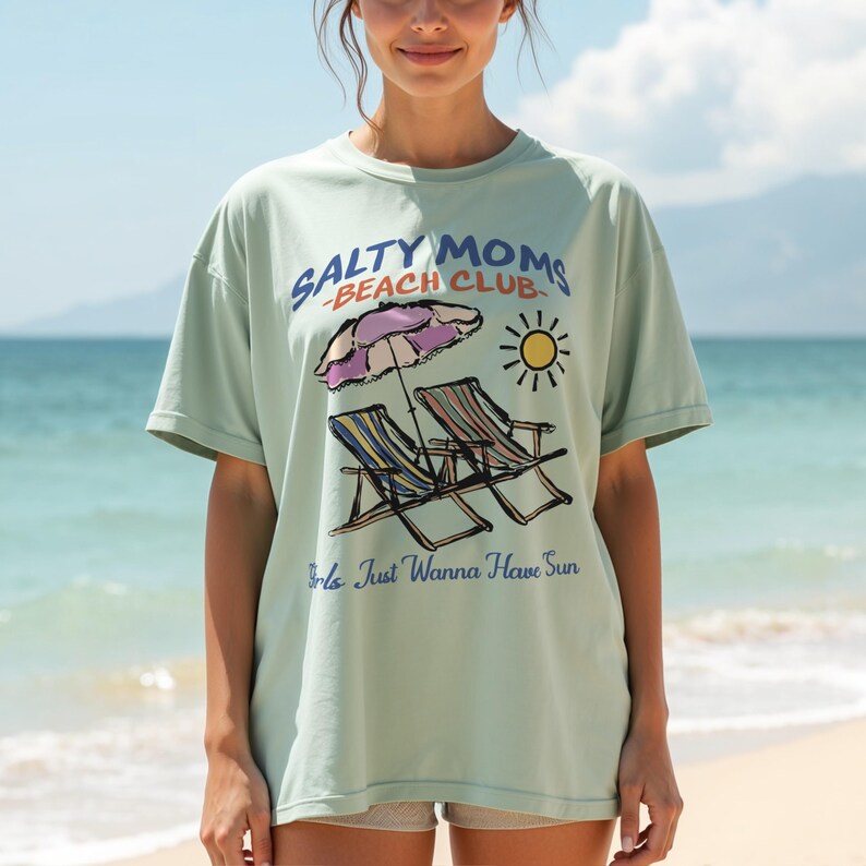 A woman is standing on a beach wearing an oversized Comfort Colors t-shirt with a design featuring a colorful hand-drawn scene of a striped beach umbrella and two chairs with the phrase Salty Moms Beach Club at the top and Girls Just Wanna Have Fun at the bottom of the design.