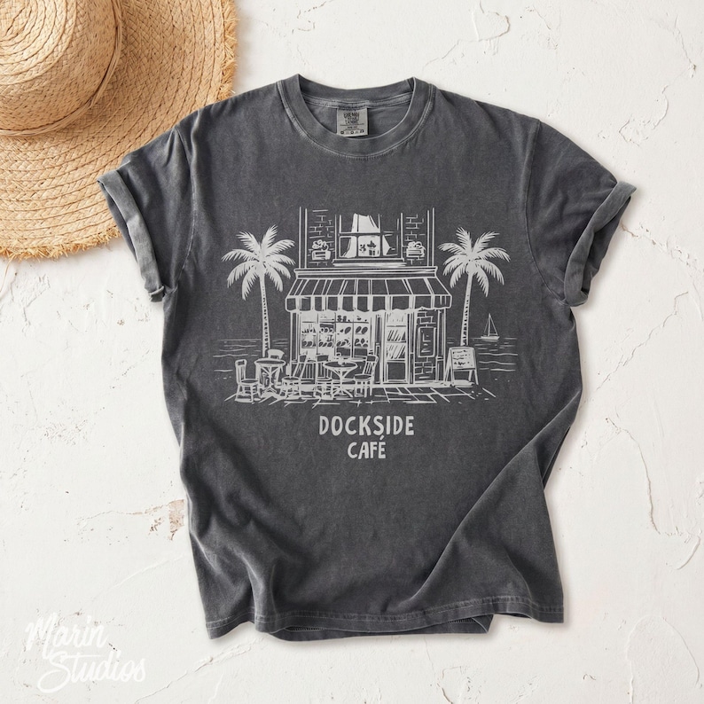Peut inclure: T-shirt gris fonc&eacute; avec une illustration blanche d'un caf&eacute;, de palmiers et le texte "Dockside Cafe". Le t-shirt a des manches retrouss&eacute;es. Un chapeau de paille est visible dans le coin sup&eacute;rieur gauche.
