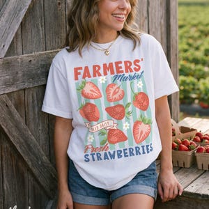 May include: White t-shirt with a "Farmers' Market" design featuring red strawberries and the text "Fresh Strawberries." The shirt is worn with denim shorts. A wooden door and a table with strawberry baskets are in the background.