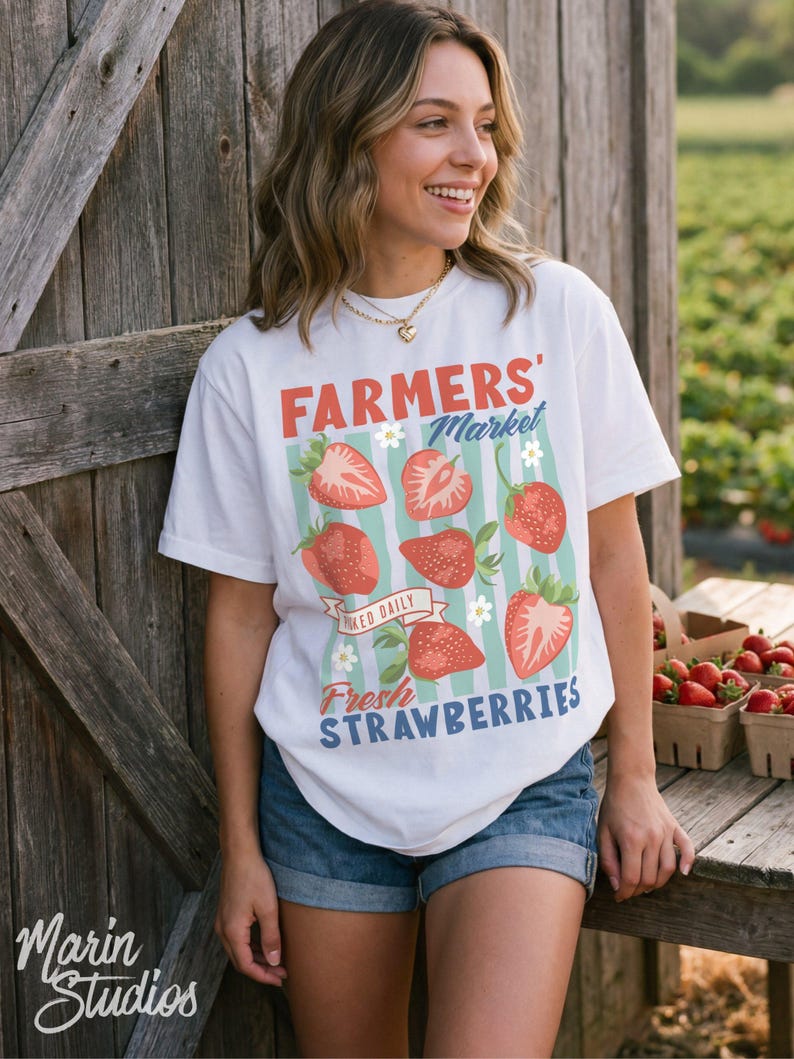 May include: White t-shirt with a "Farmers' Market" design featuring red strawberries and the text "Fresh Strawberries." The shirt is worn with denim shorts. A wooden door and a table with strawberry baskets are in the background.