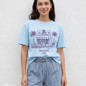 Peut inclure: T-shirt bleu clair avec un dessin de caf&eacute;, de palmiers et le texte "Dockside Cafe". Le mod&egrave;le porte un short en jean. L'image est prise devant un mur blanc.