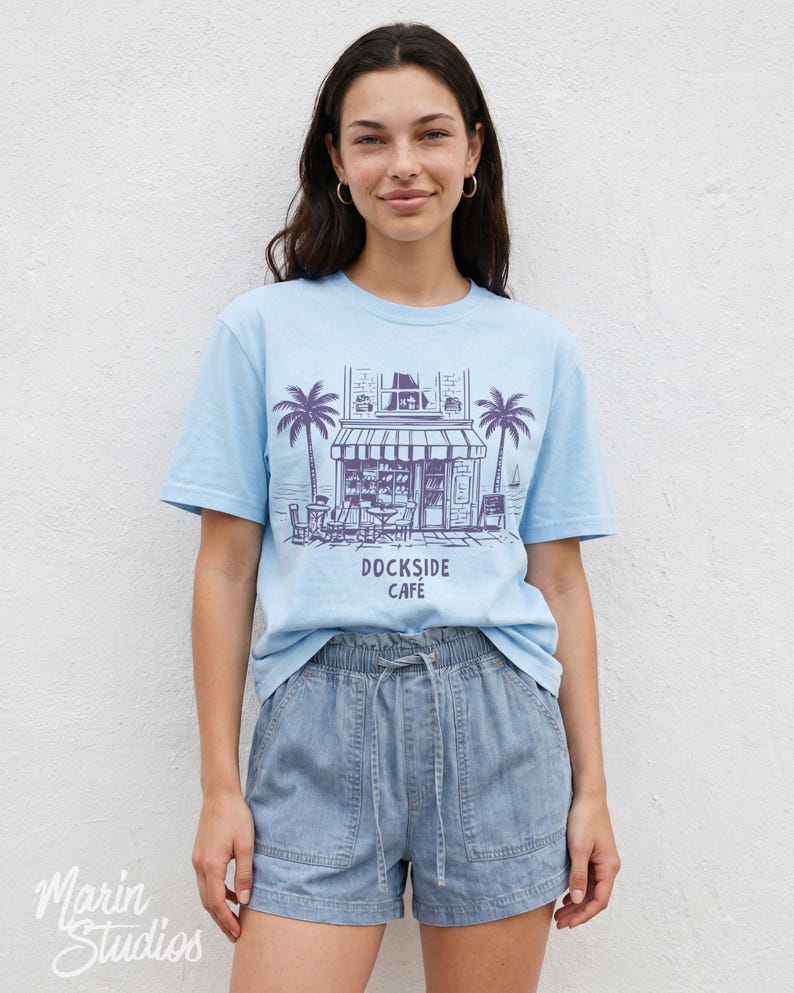 Peut inclure: T-shirt bleu clair avec un dessin de caf&eacute;, de palmiers et le texte "Dockside Cafe". Le mod&egrave;le porte un short en jean. L'image est prise devant un mur blanc.