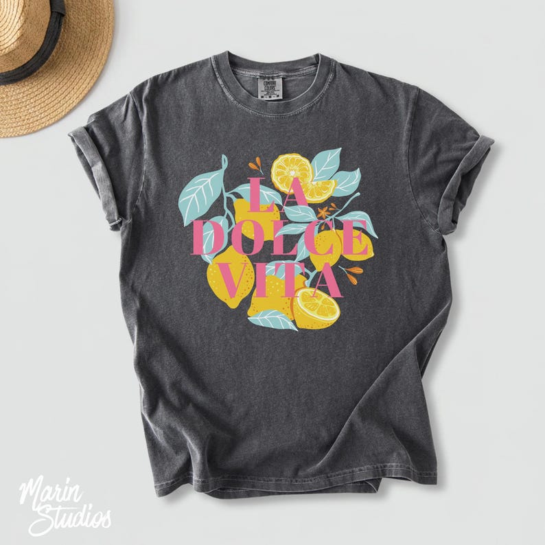 May include: Dark gray t-shirt featuring a colorful graphic of lemons and leaves with the text "LA DOLCE VITA" in pink and light blue. The shirt has a vintage, washed look.