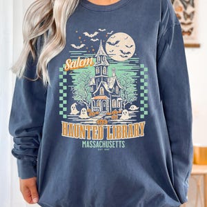 A woman is wearing an oversized Denim Comfort Colors sweatshirt with a design featuring a haunted library with ghosts and pumpkins and the phrase Salem Haunted Library, Massachusetts.