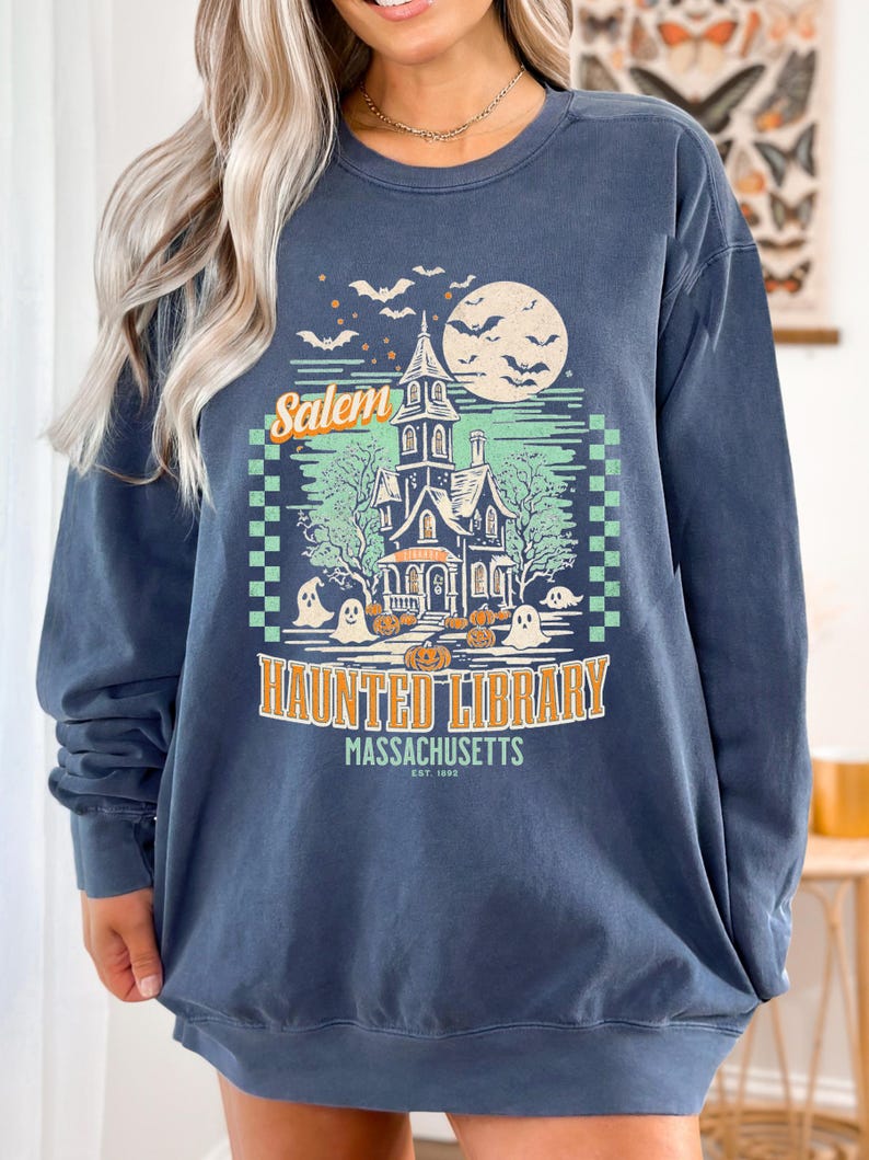 A woman is wearing an oversized Denim Comfort Colors sweatshirt with a design featuring a haunted library with ghosts and pumpkins and the phrase Salem Haunted Library, Massachusetts.