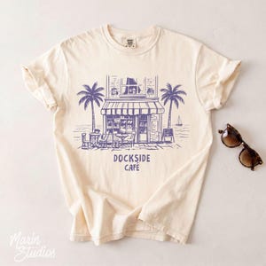 Peut inclure: T-shirt cr&egrave;me avec une illustration bleue d'un caf&eacute; avec des palmiers, des tables et des chaises. Le texte "DOCKSIDE CAFE" est imprim&eacute; en dessous. Des lunettes de soleil sont pos&eacute;es sur le c&ocirc;t&eacute; droit du t-shirt.