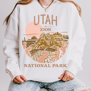 Woman wearing a White oversized sweatshirt with a vintage graphic of Zion National Park in Utah, featuring canyons, cacti, and a sunset design, styled with ripped jeans.