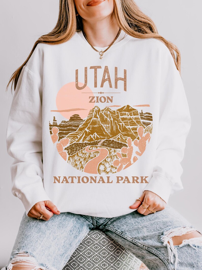 Woman wearing a White oversized sweatshirt with a vintage graphic of Zion National Park in Utah, featuring canyons, cacti, and a sunset design, styled with ripped jeans.