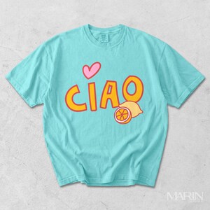 K&ouml;nnte beinhalten: Ein t&uuml;rkisfarbenes T-Shirt mit dem Wort "CIAO" in orangefarbenen Buchstaben mit gelber Umrandung, einem rosa Herz und einer Zitronenillustration. Das Shirt besteht aus weichem Material und hat kurze &Auml;rmel.
